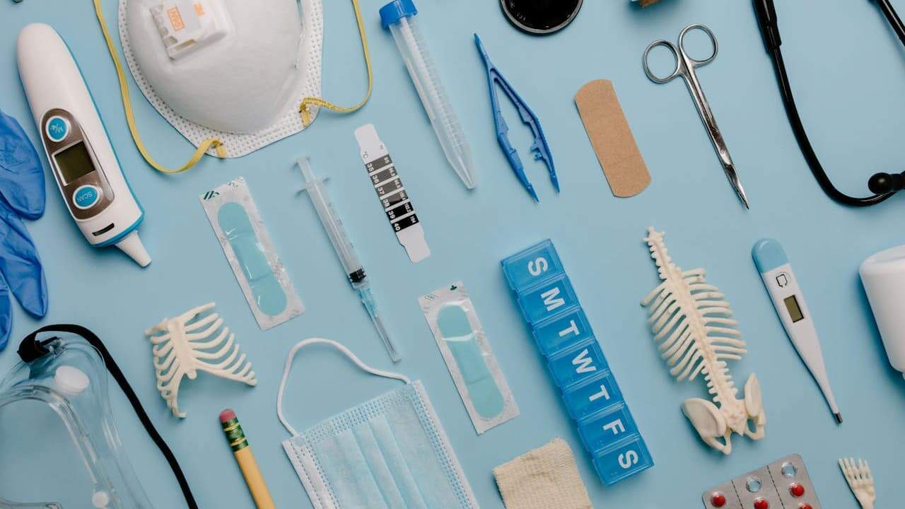Medical supplies and equipment arranged on blue surface including masks, thermometer, syringes, bandages, and skeleton models
