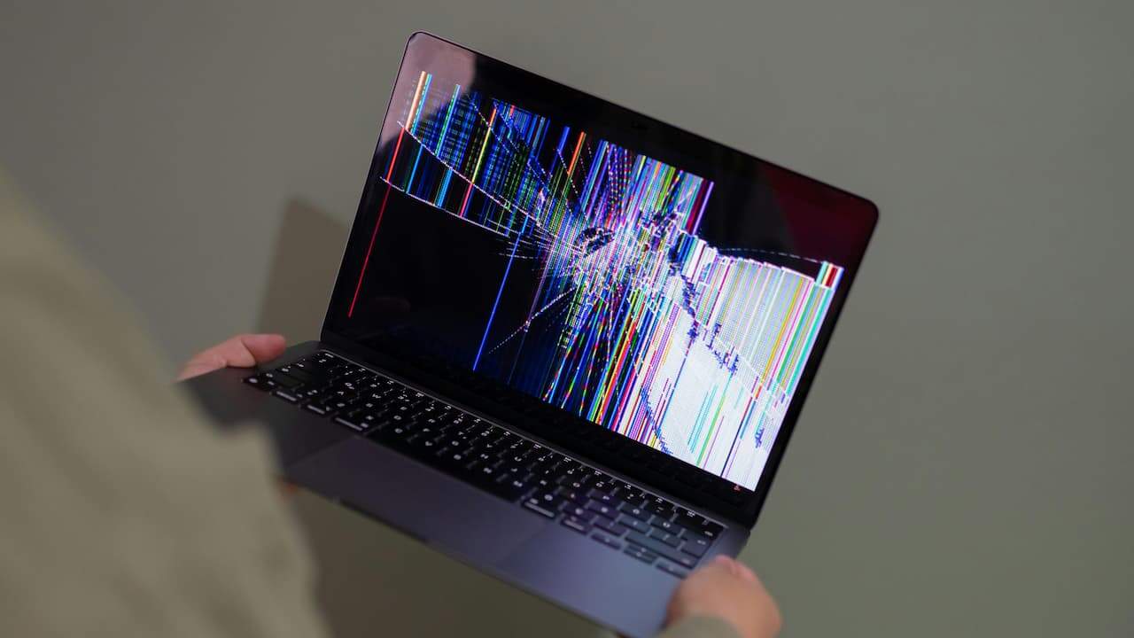 Person holding a laptop with a severely cracked and distorted screen displaying multicolored vertical and horizontal lines and black spots from impact damage