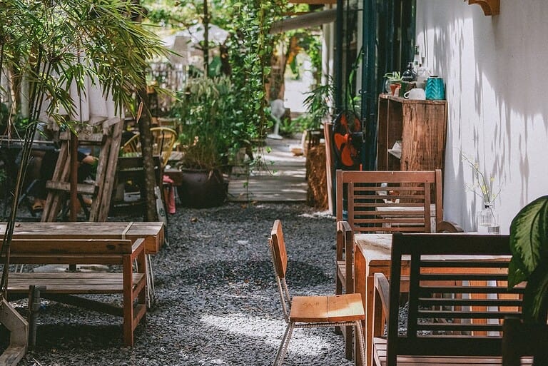 Outdoor seating area with wooden chairs and tables in courtyard, shaded garden restaurant space, rustic dining furniture on stone paving, empty cafe patio with plants and greenery, charming restaurant exterior with natural setting