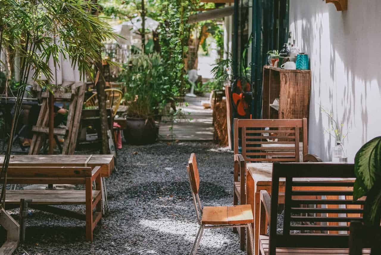 Outdoor seating area with wooden chairs and tables in courtyard, shaded garden restaurant space, rustic dining furniture on stone paving, empty cafe patio with plants and greenery, charming restaurant exterior with natural setting
