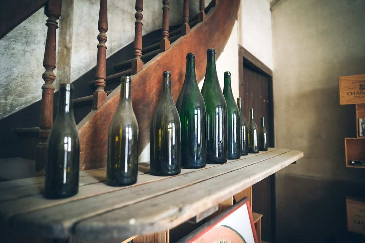 Vintage green glass wine bottles lined up on rustic wooden shelf near ornate wooden staircase with turned balusters