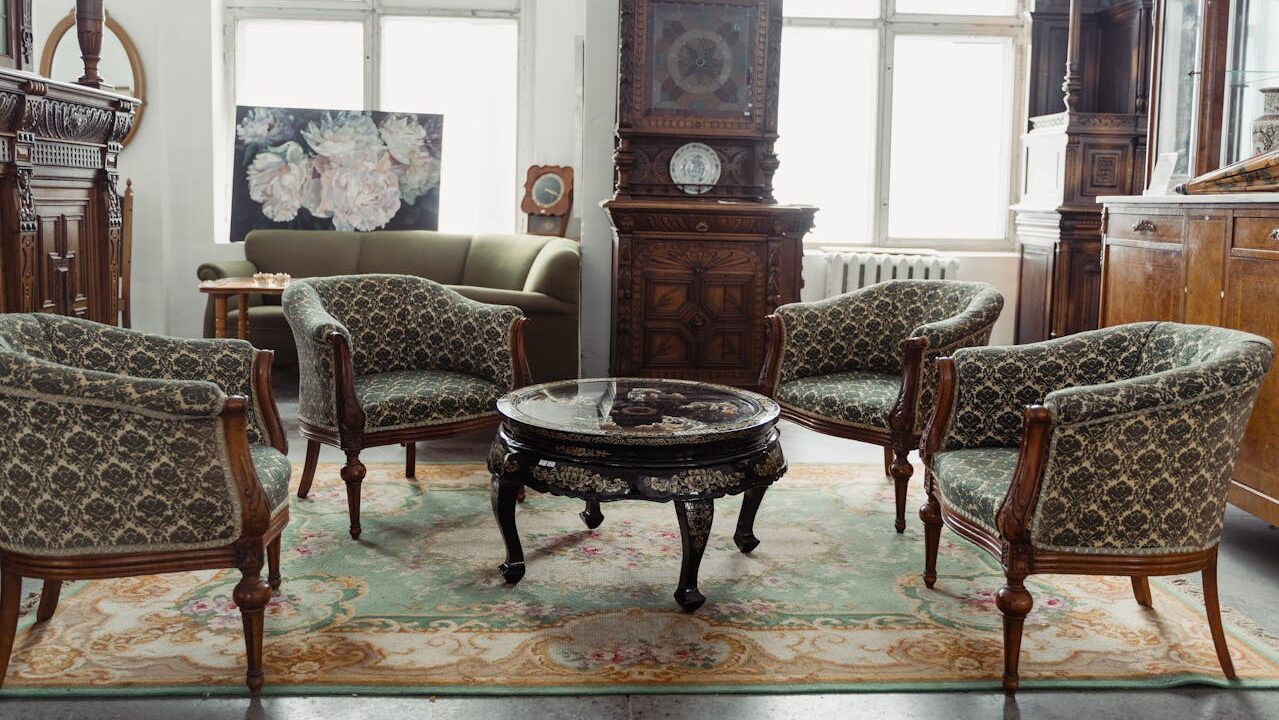 Elegant vintage living room with patterned armchairs around ornate coffee table, antique cabinet, floral rug, and tall windows