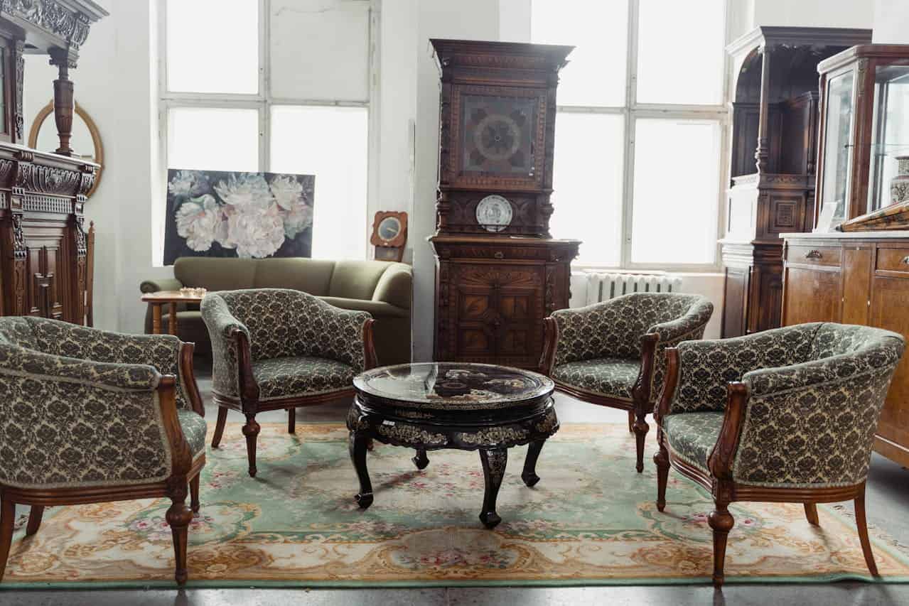 Elegant vintage living room with patterned armchairs around ornate coffee table, antique cabinet, floral rug, and tall windows