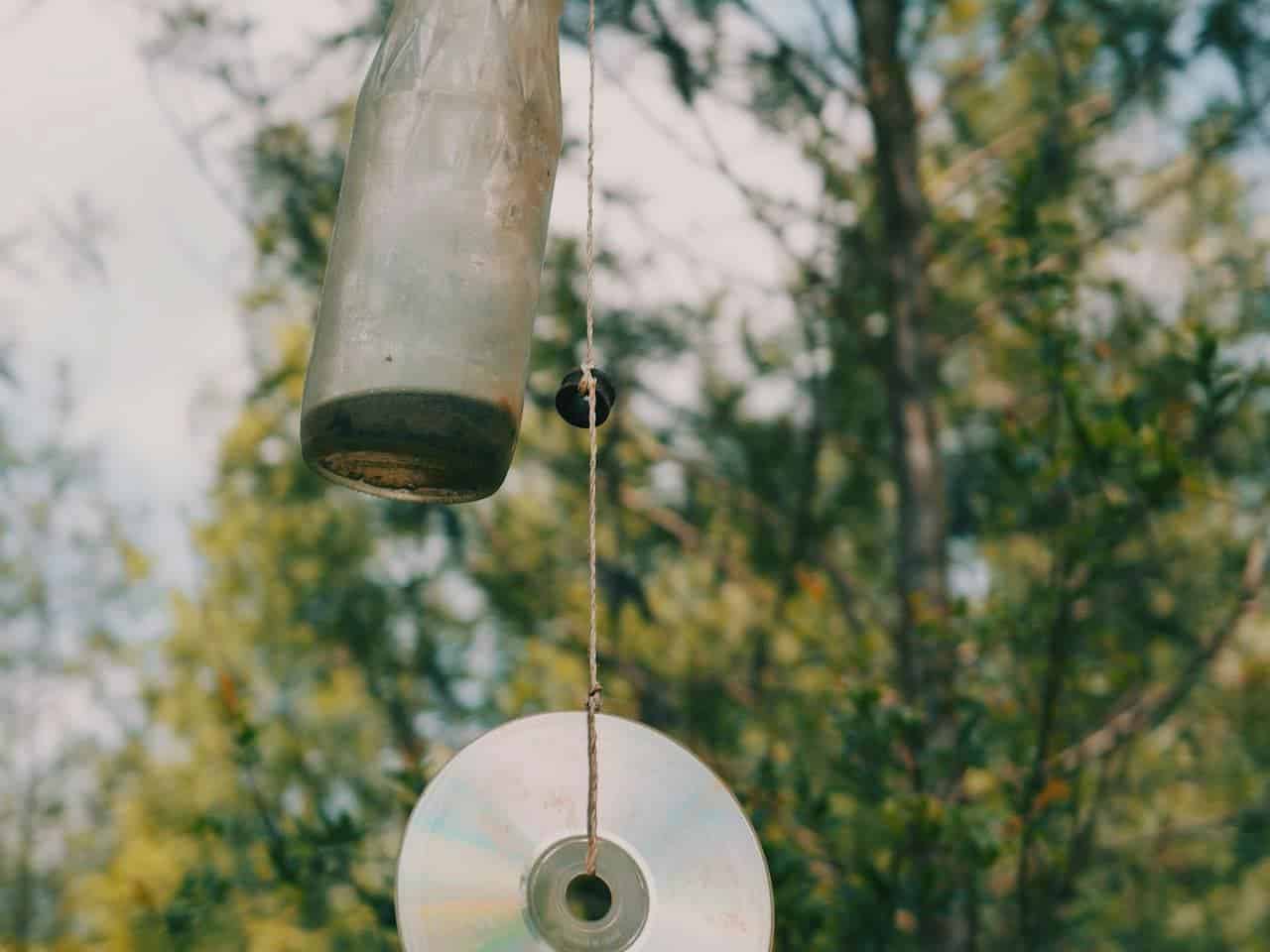 A plastic bottle and a compact disc (CD) hanging side by side from strings or wires, suspended against a blurred natural background
