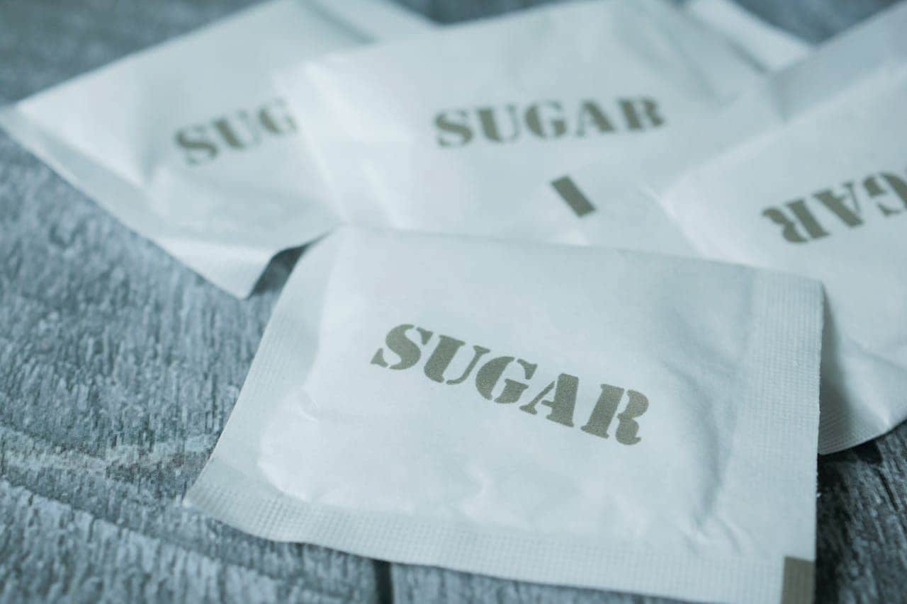 Several white sugar packets labeled "SUGAR" scattered on a gray wooden surface, with one packet prominently displayed in foreground