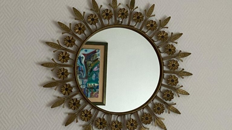 Round mirror with decorative brass sunburst frame featuring flower and leaf details, mounted on textured white wall