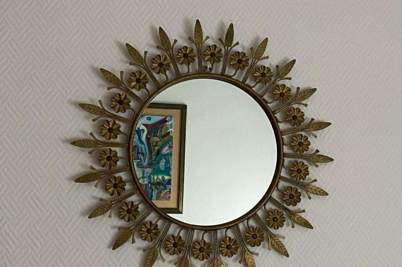 Round mirror with decorative brass sunburst frame featuring flower and leaf details, mounted on textured white wall