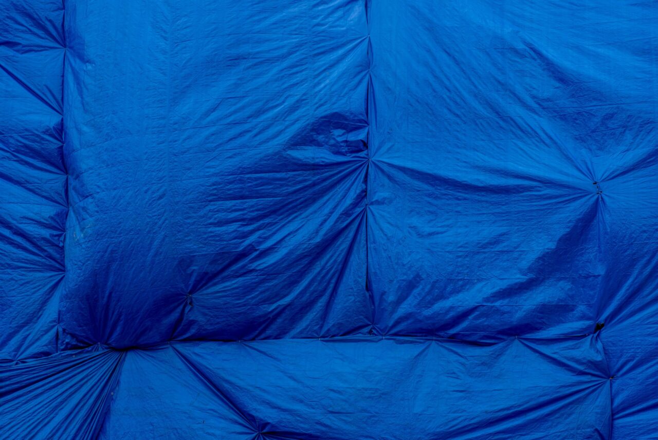 Blue silage tarp, crinkled surface, tightly pinned at intervals, durable waterproof material, used for agricultural coverage, slightly bulging areas indicating underlying content