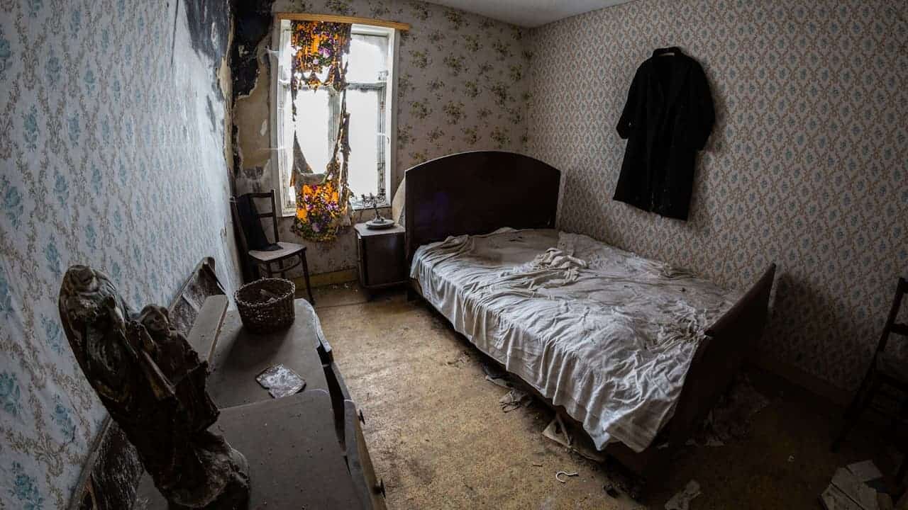 Dimly lit bedroom, single bed with patterned blanket, vintage wooden furniture, floral wallpaper, clothes hanging on wall, curtain partially open, small and modest room, cozy but slightly cluttered