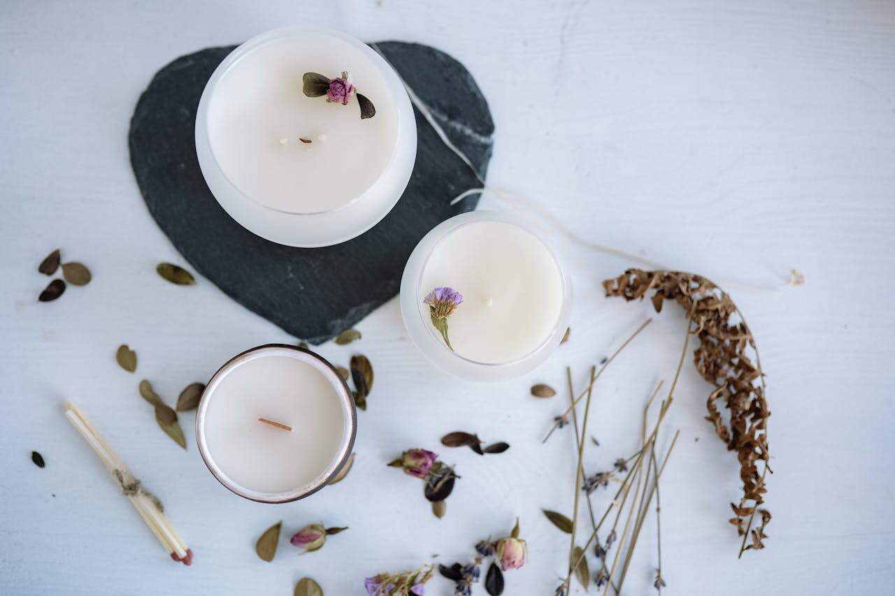 Three white scented candles with lavender on marble surface, decorated with dried flowers. Heart-shaped slate coaster and scattered botanical elements surrounding them
