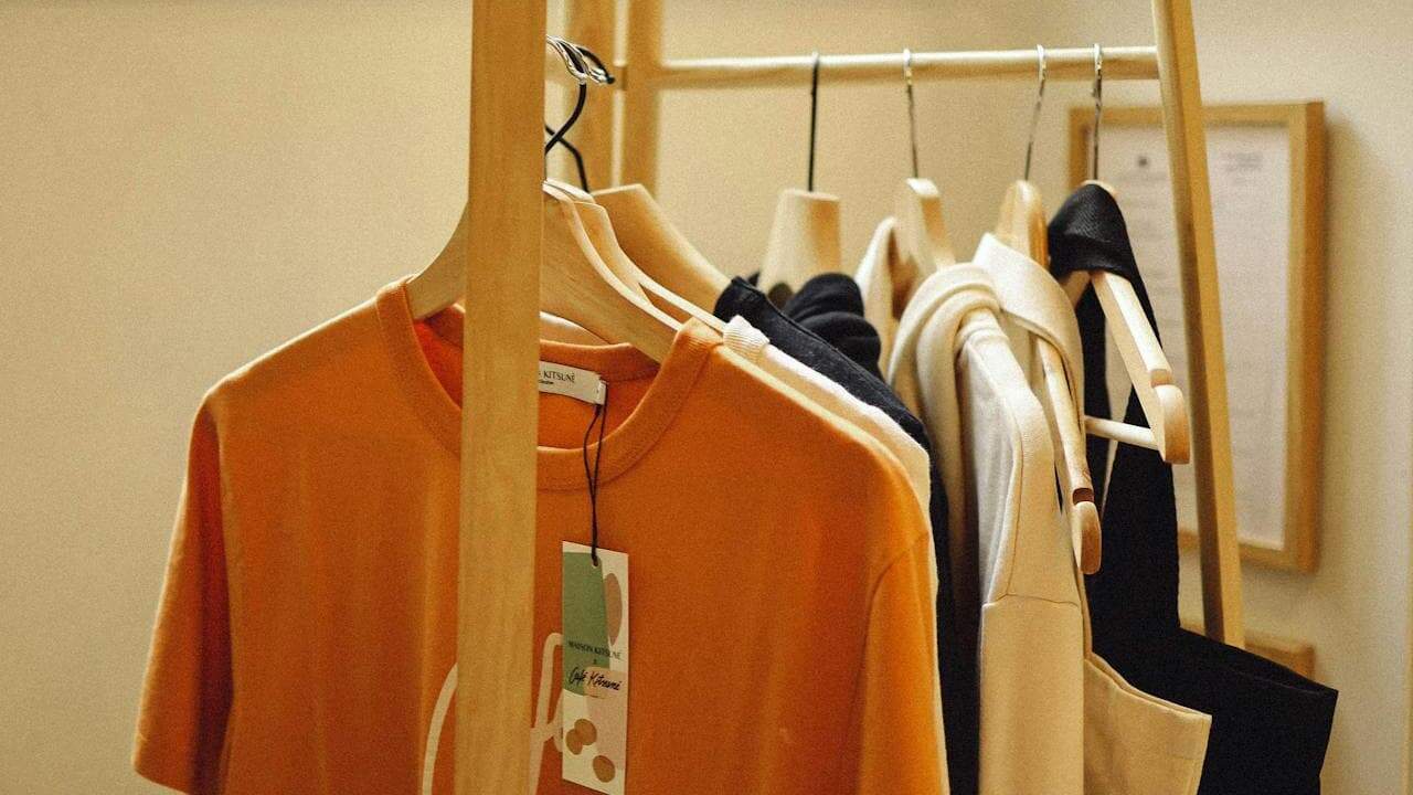Clothing rack with a variety of neatly hung garments, including an orange over stetched t-shirt with a visible tag, and neutral-toned jackets and tops