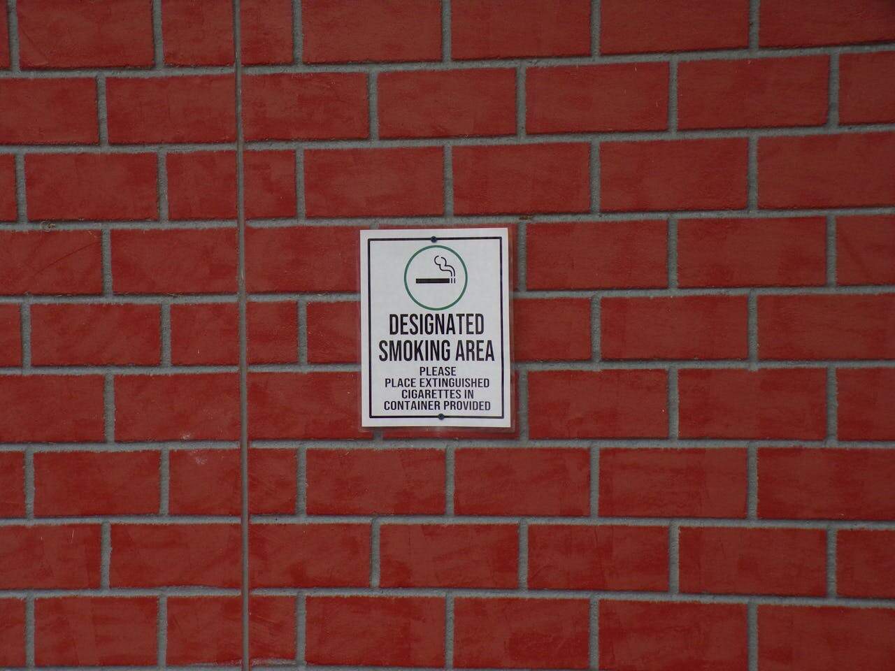 Sign reading “Designated Smoking Area” on a red brick wall, black text with smoking icon, instructing cigarette disposal in provided container