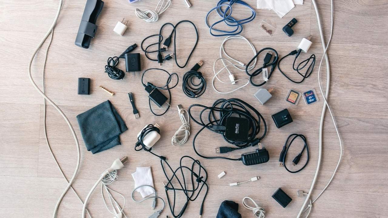 Assorted electronic cables, chargers, adapters, memory cards, and plugs scattered randomly on a light wooden surface