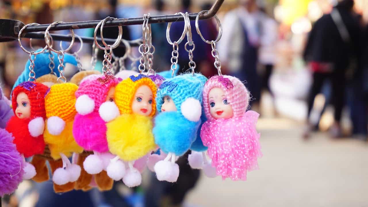 Colorful doll-faced keychains in knitted outfits hanging on a metal rack, background shows a blurred outdoor market scene