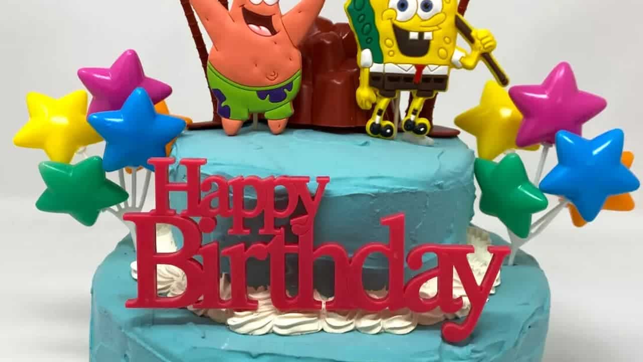 Blue two-tier birthday cake with SpongeBob and Patrick figurines on top, colorful star decorations and red "Happy Birthday" text