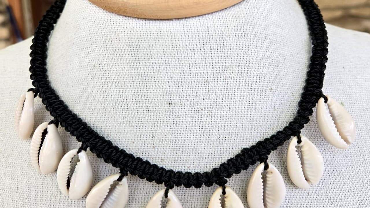 Black braided rope necklace with white cowrie shells displayed on white fabric mannequin bust against wooden background