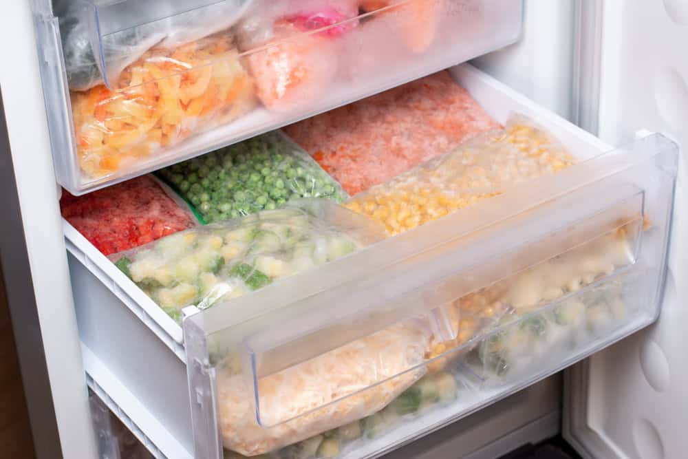 Open freezer drawer containing clear plastic bags of various frozen vegetables and fruits organized in compartments