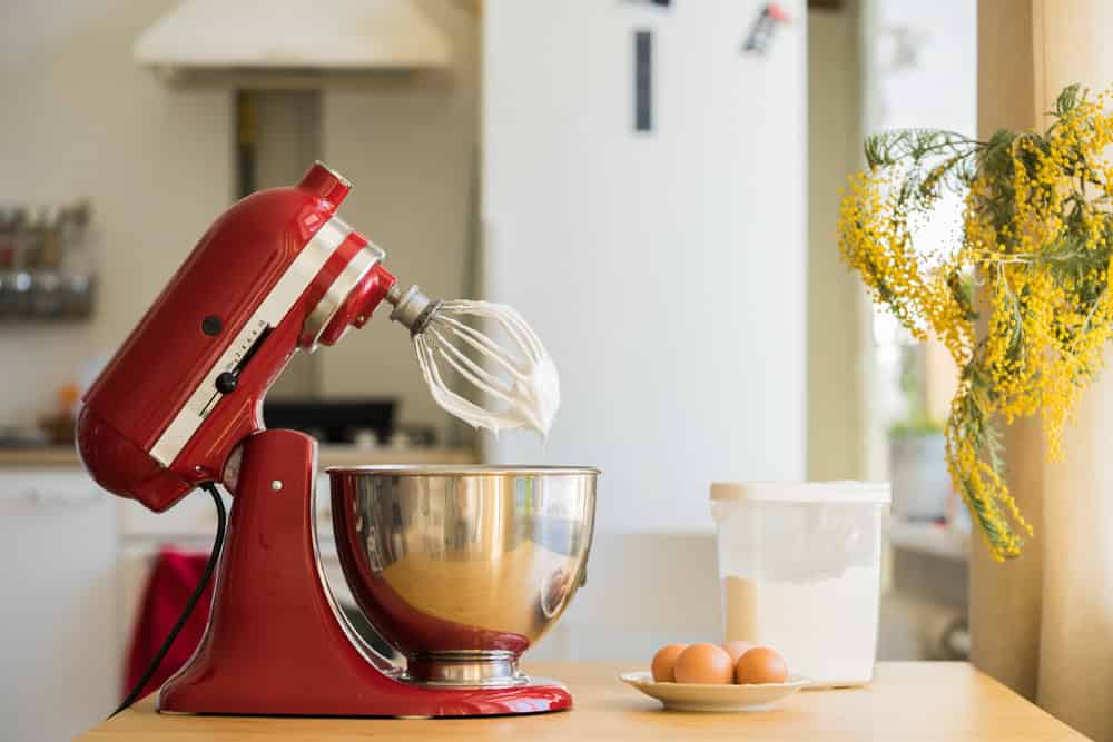 A red stand mixer with a whisk attachment and stainless steel bowl on a kitchen counter, next to a container of flour, a plate with three eggs