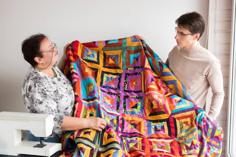 Two women displaying colorful patchwork quilt with geometric squares and vibrant fabric patterns