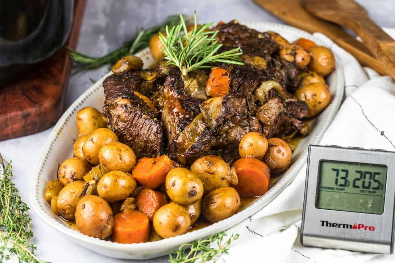 Cooked pot roast served on a white platter, surrounded by baby potatoes and carrots, garnished with rosemary, with a digital meat thermometer reading 205°F placed beside the dish