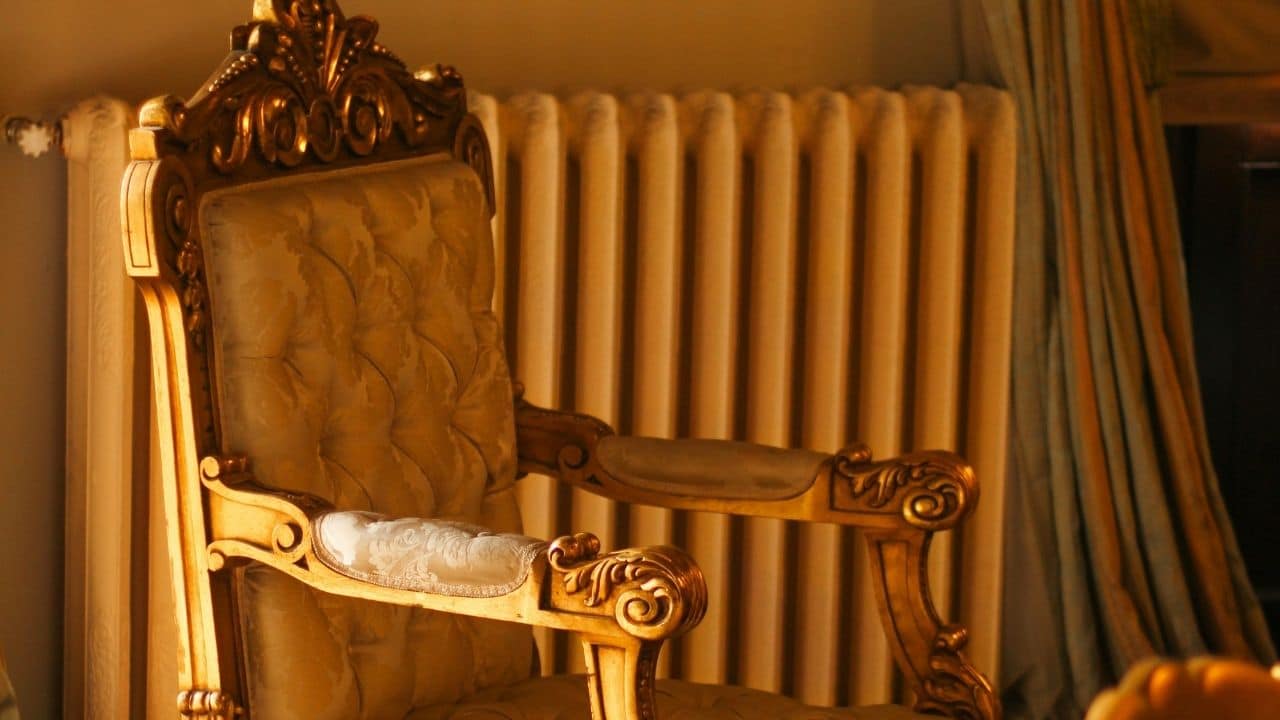 ornate vintage armchair, carved wooden armrests, tufted golden upholstery, placed in front of a cream radiator, warm indoor lighting, classic interior décor