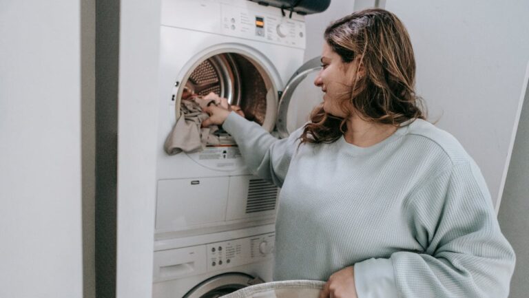 Woman with long hair wearing light blue sweater loading clothes into a stacked washer-dryer unit, holding laundry basket