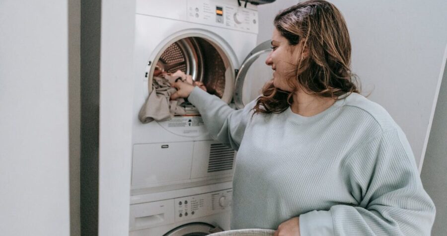 Woman with long hair wearing light blue sweater loading clothes into a stacked washer-dryer unit, holding laundry basket
