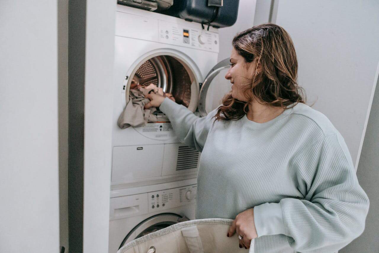 Woman with long hair wearing light blue sweater loading clothes into a stacked washer-dryer unit, holding laundry basket
