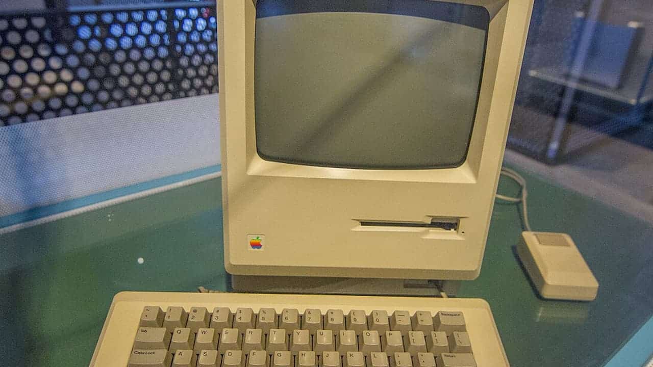 Vintage beige Apple Macintosh computer with CRT monitor, keyboard, and mouse displayed on green glass table surface