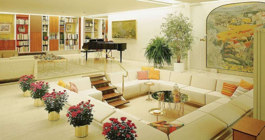 Sunken living room with white sectional sofa, grand piano, built-in bookshelves, and red flowering plants in golden planters