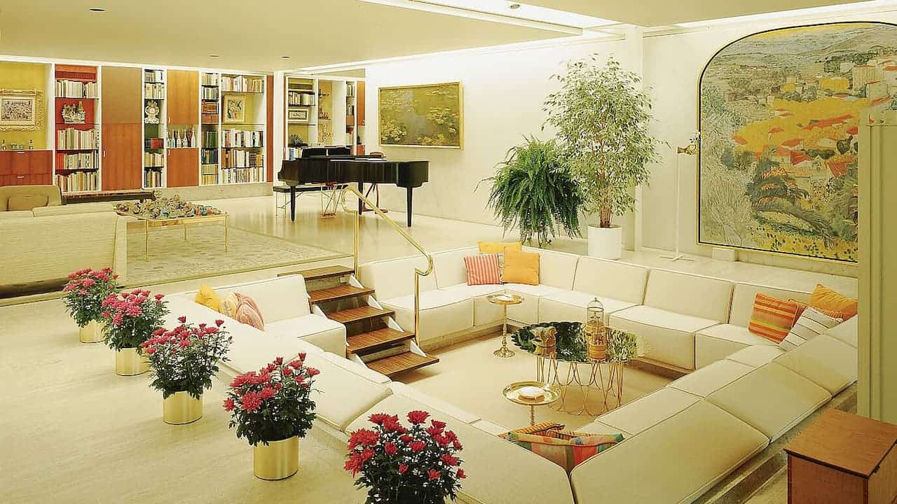 Sunken living room with white sectional sofa, grand piano, built-in bookshelves, and red flowering plants in golden planters