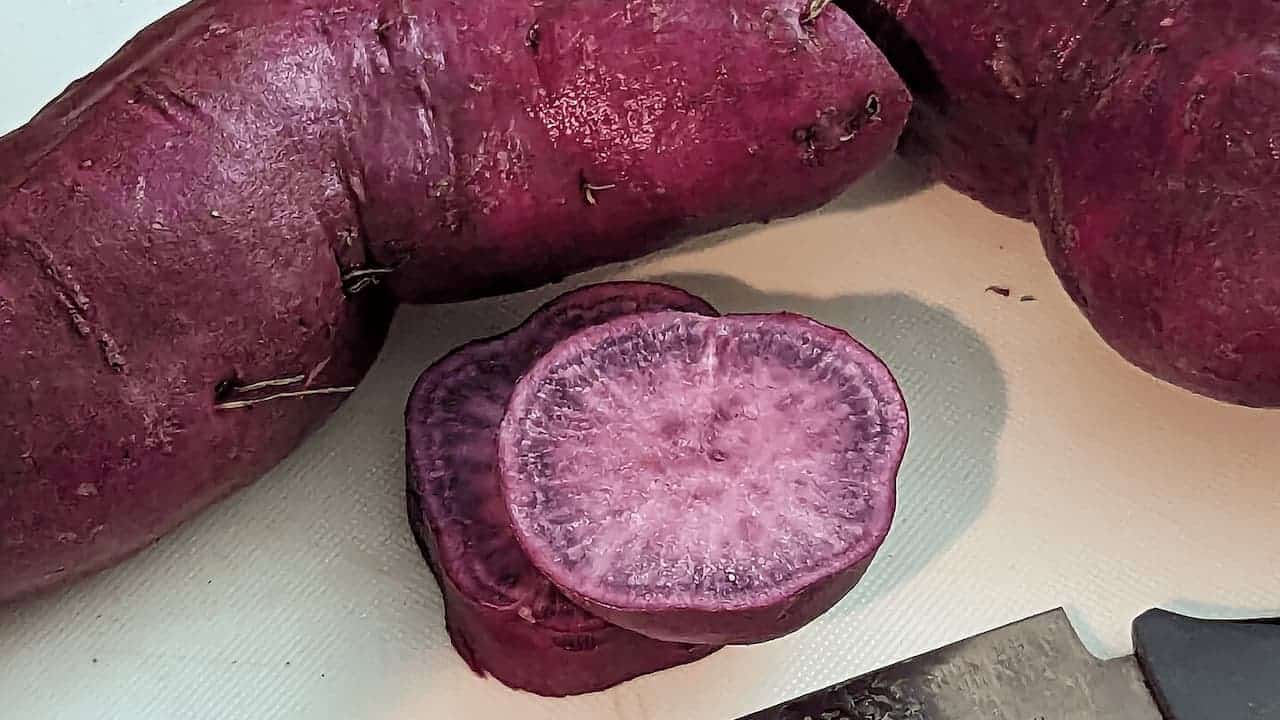 Purple sweet potatoes with one sliced to reveal vibrant purple flesh interior on white cutting board surface