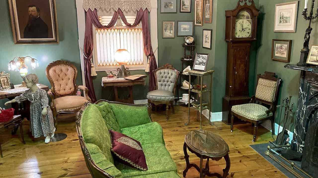 Victorian parlor room with green velvet furniture, grandfather clock, stained glass window, portrait paintings, and hardwood floors