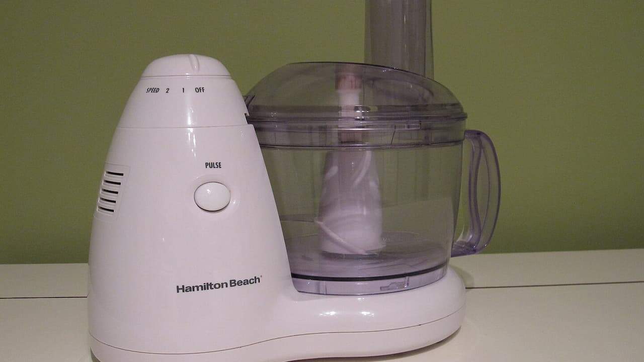 White Hamilton Beach food processor with clear bowl, speed controls, pulse button on white counter against green wall