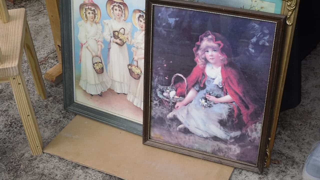 Two framed vintage oil paintings featuring women in period dress with flower baskets, displayed on concrete floor near wooden easel
