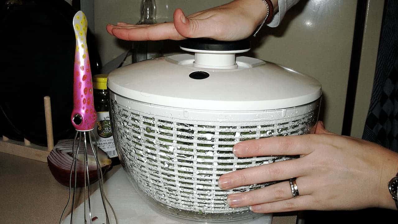 Person operating white salad spinner with perforated basket, hands positioned on lid and base in kitchen setting