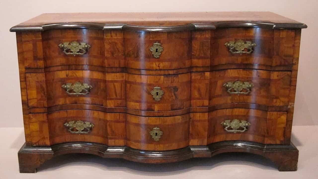 Antique wooden chest of drawers with curved front, ornate brass hardware, and rich brown finish against neutral wall
