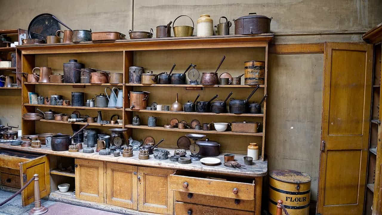 Historic butler's pantry with wooden shelving displaying extensive collection of copper pots, ceramic crocks, baking molds, and kitchenware