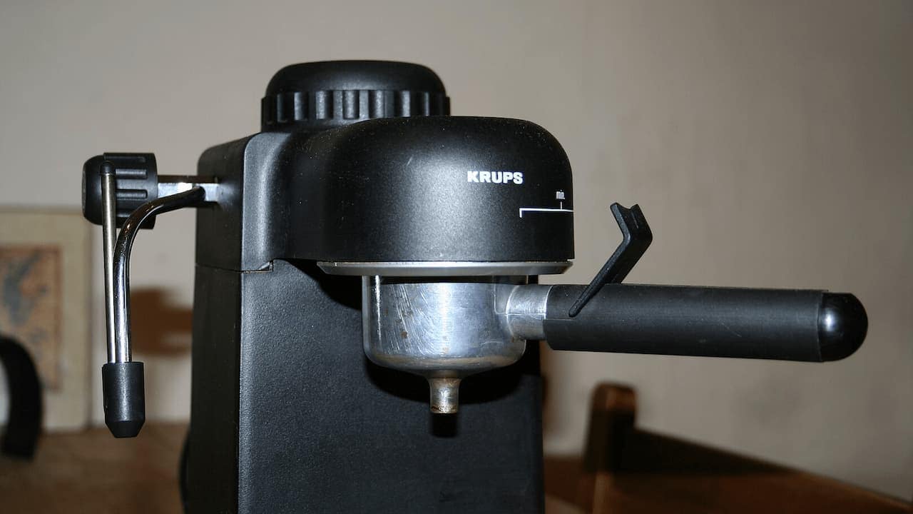 Black Krups espresso machine with steam wand and portafilter on counter against beige wall with framed artwork