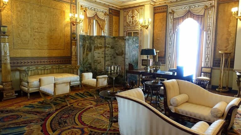 Ornate historic fainting room with gold silk walls, cream upholstered furniture, decorative fireplace screen, crystal chandeliers, elaborate moldings