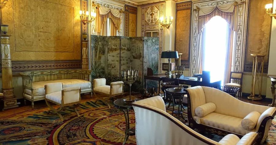 Ornate historic fainting room with gold silk walls, cream upholstered furniture, decorative fireplace screen, crystal chandeliers, elaborate moldings