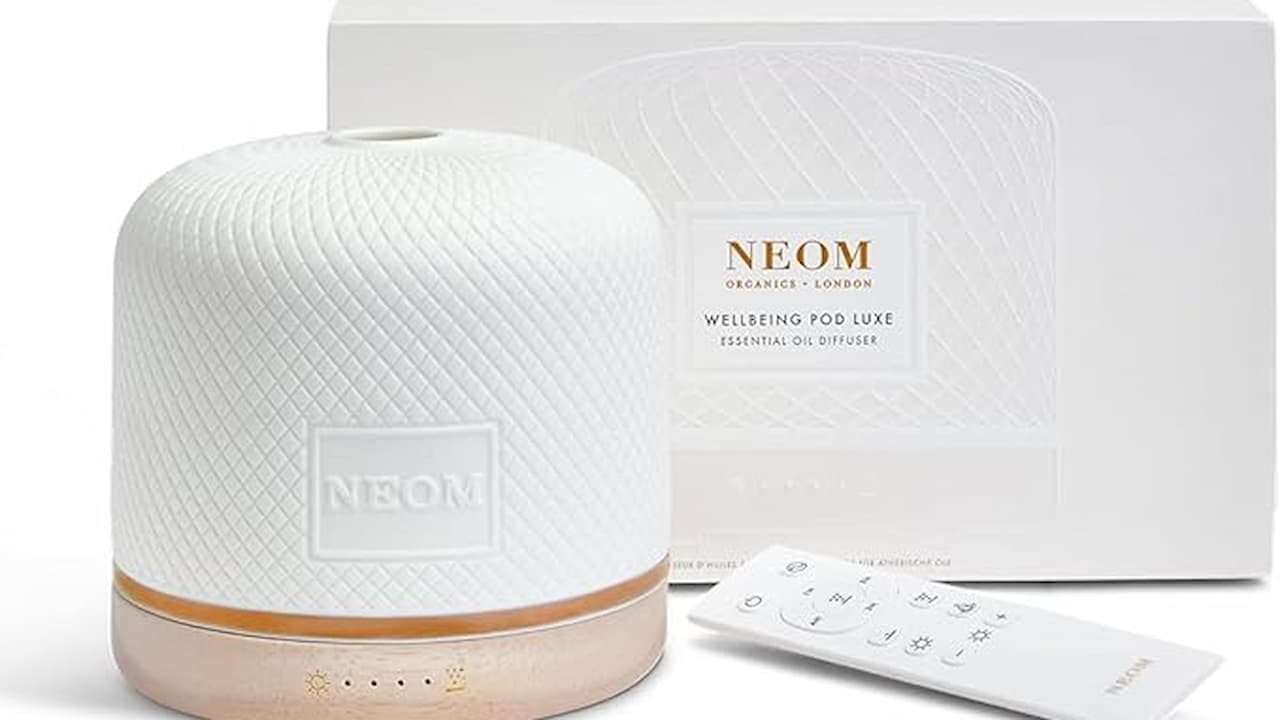 White NEOM Wellbeing Pod Luxe essential oil diffuser with a textured surface, wooden base, and matching white remote control, displayed in front of its branded packaging box