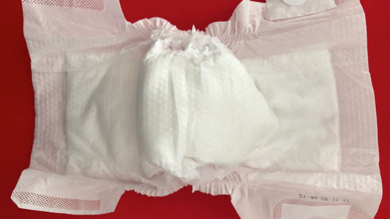 Disposable baby diaper laid open on a red surface, white padded center with side flaps and elastic leg cuffs, textured absorbent material visible