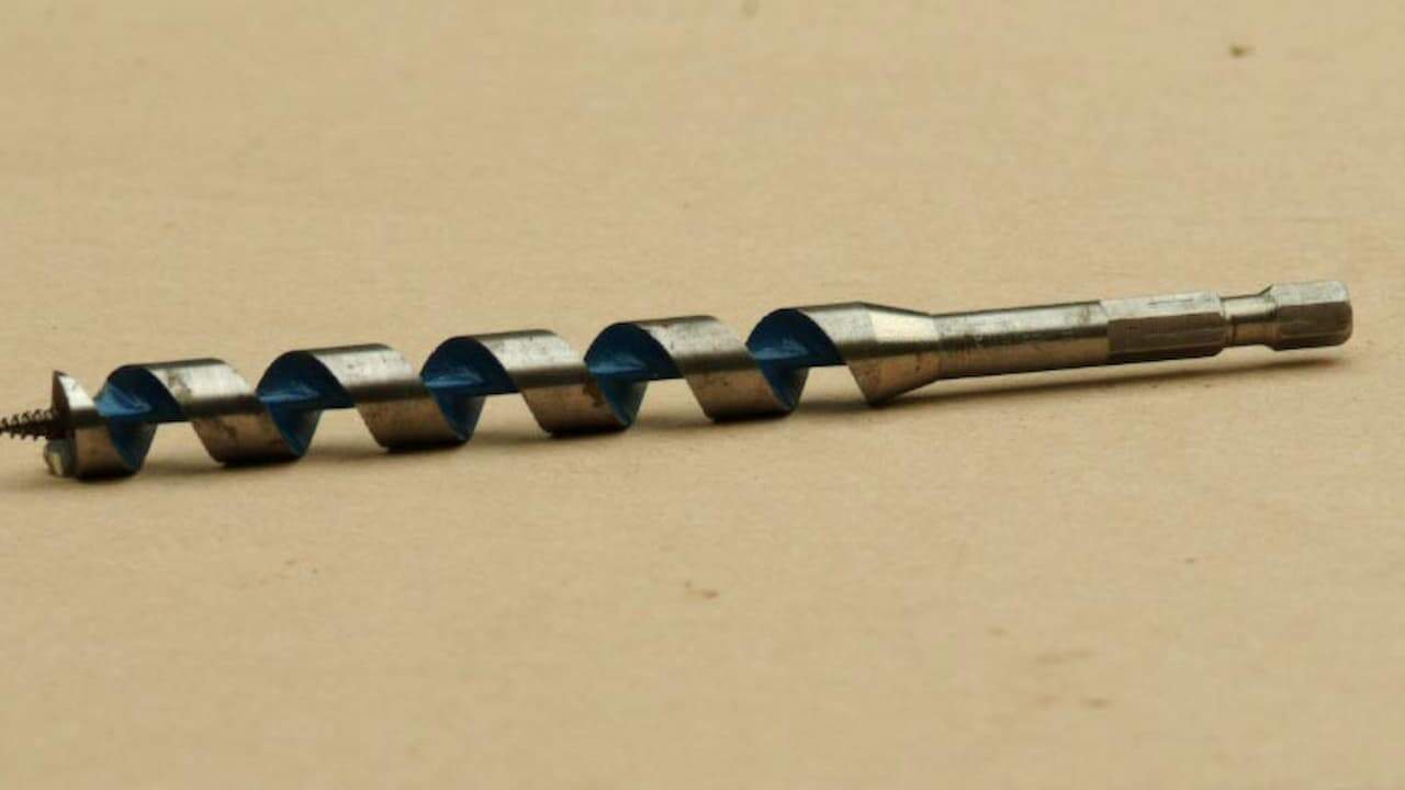 Metal auger drill bit with spiral flutes and hexagonal shank lying on light wooden surface for drilling holes