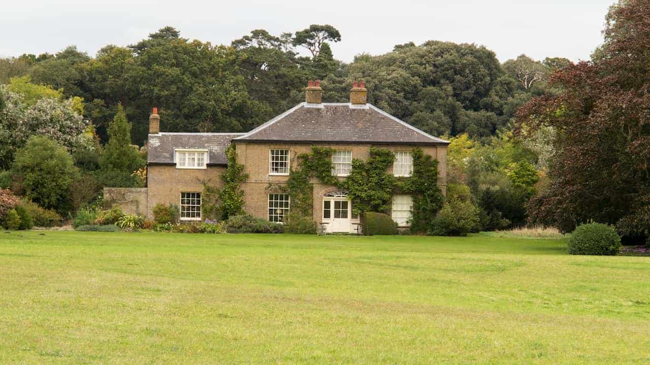 Large countryside cottage, ivy-covered brick walls, symmetrical windows with white frames, twin chimneys on pitched roof, wide green lawn in foreground