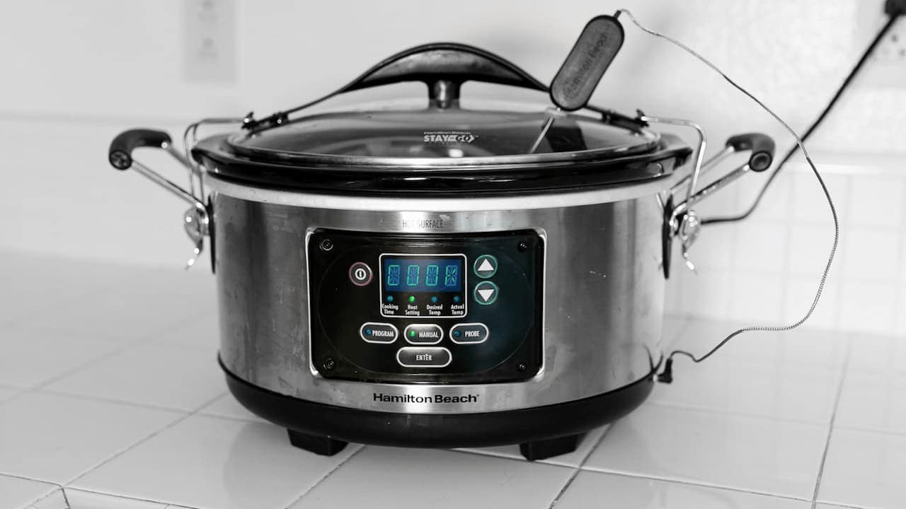 Hamilton Beach slow cooker with digital display, stainless steel finish, glass lid, and carrying clips on tiled surface