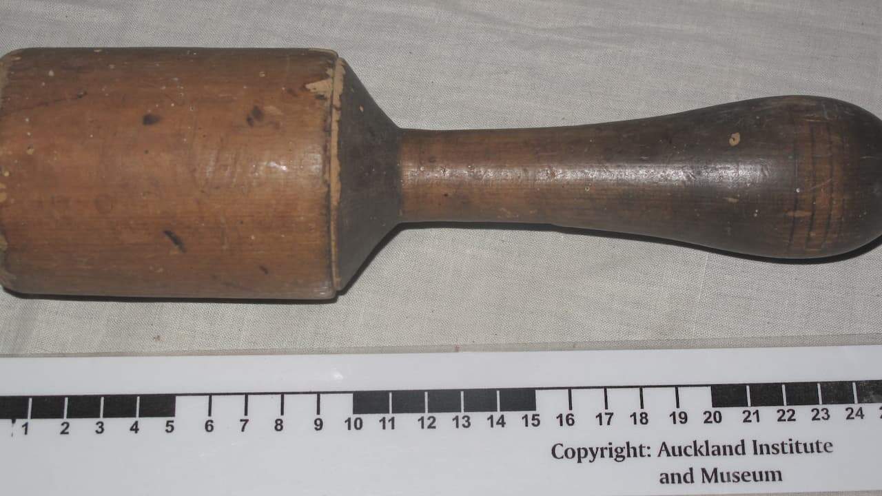 Wooden potato masher with a thick cylindrical head and tapered handle, placed on a cloth surface next to a measurement scale