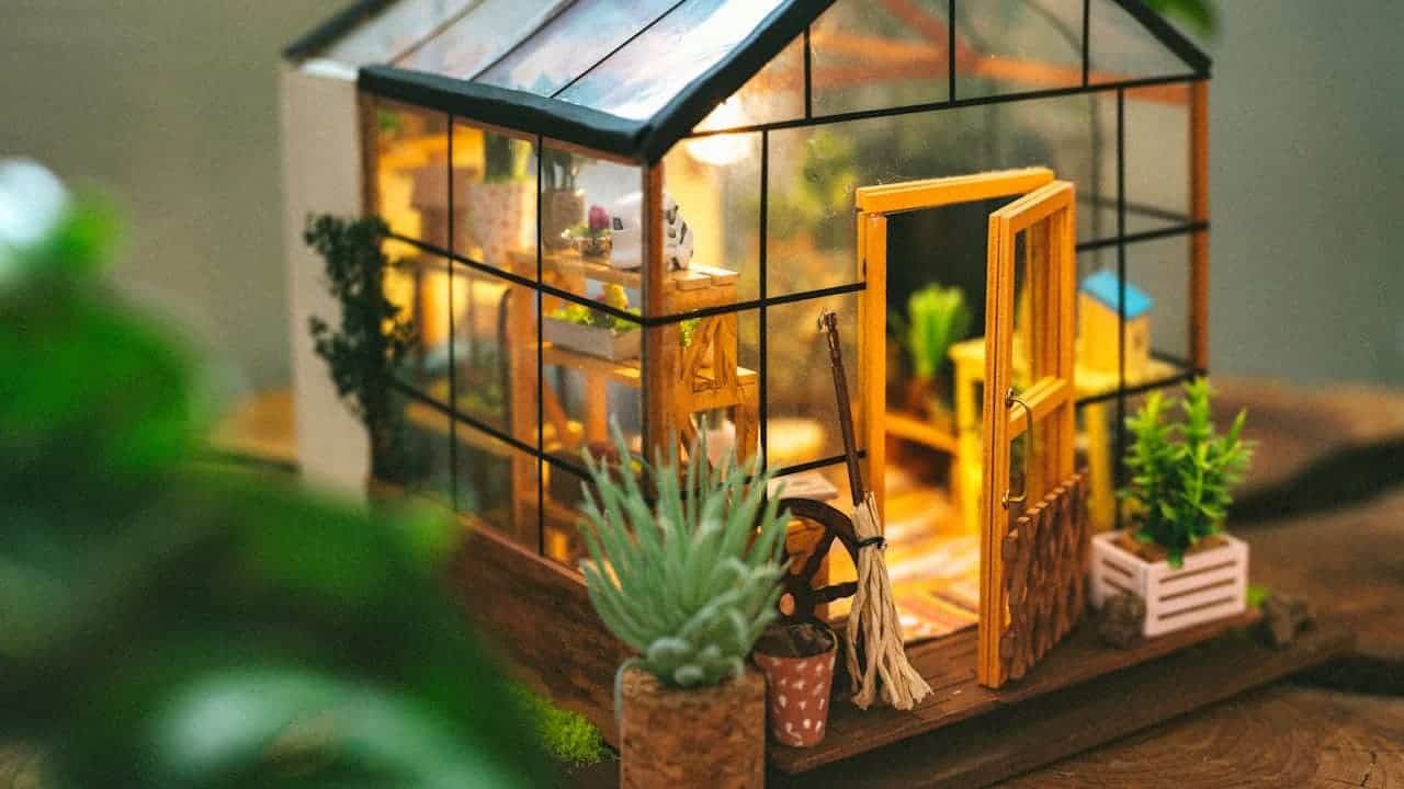Miniature glass greenhouse dollhouse with warm lighting, tiny plants, garden tools, wooden shelving, detailed interior furnishings, craft model