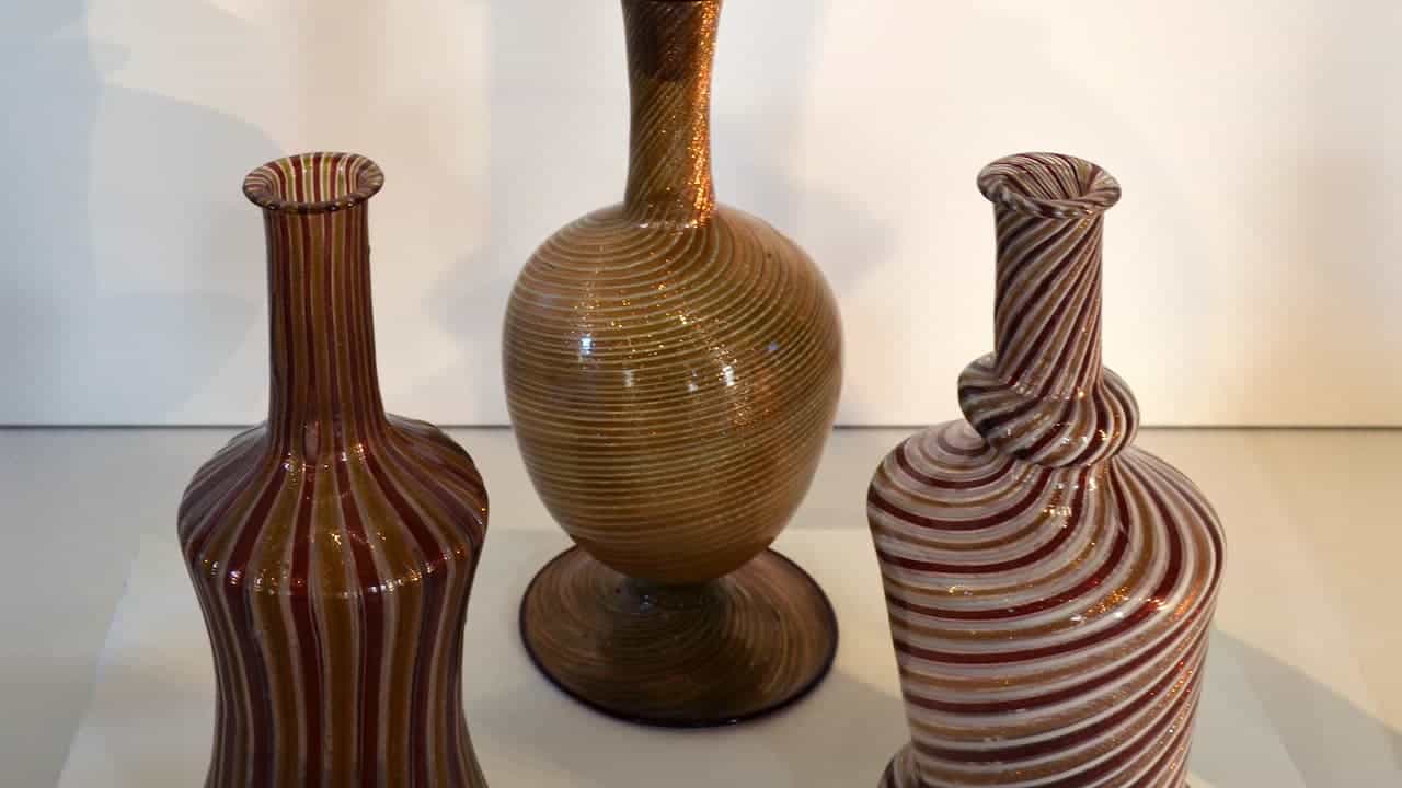 Three Murano glass vases with intricate spiral patterns in red, gold, and white hues, displayed against a neutral background