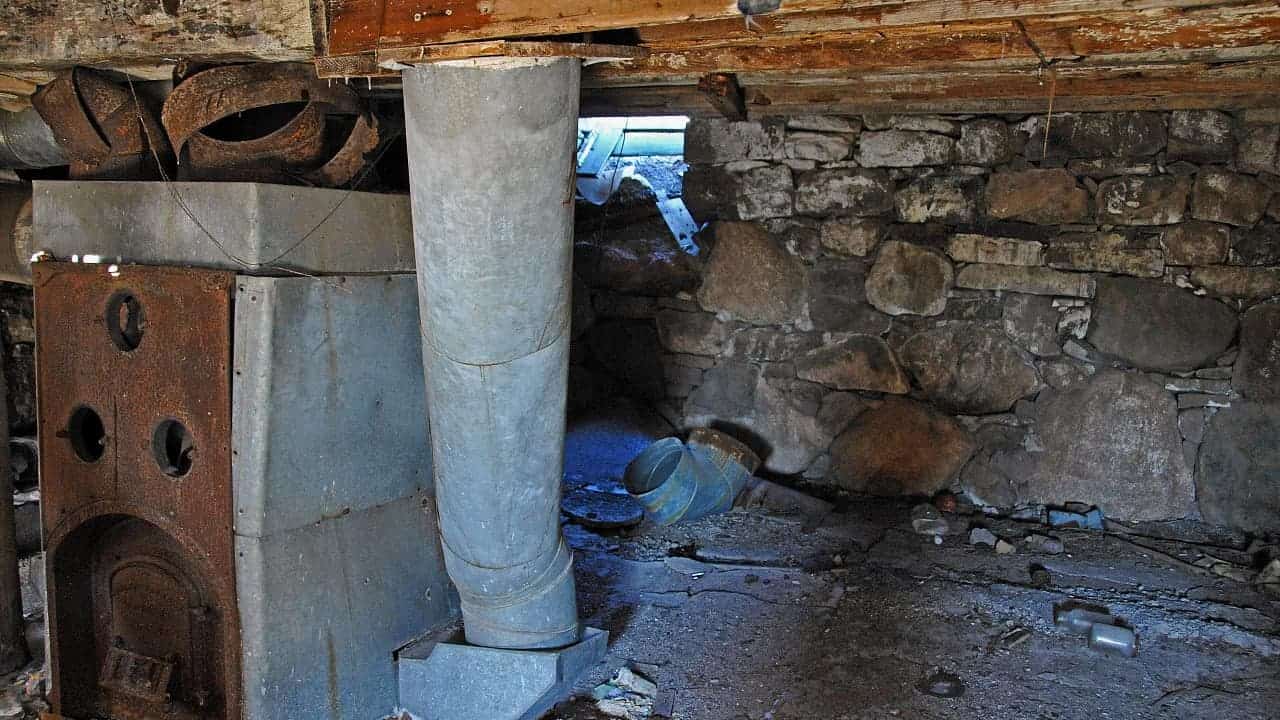 Abandoned stone basement with rusty coal furnace, metal ductwork, deteriorating walls, debris scattered on wet concrete floor